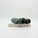 A pair of green binoculars displayed upright on a white branded stand against a clean white background.